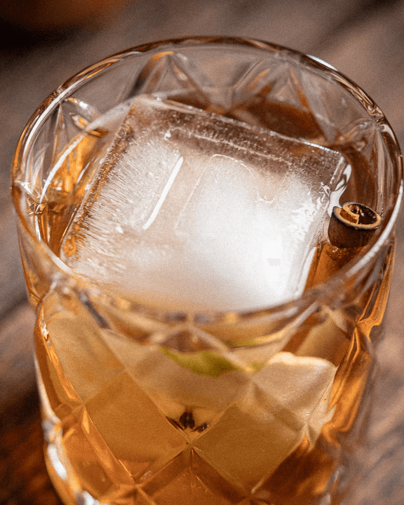 Apple Crisp cocktail in a rocks glass over one large clear cube, garnished with a thin apple slice and a charred cinnamon stick.